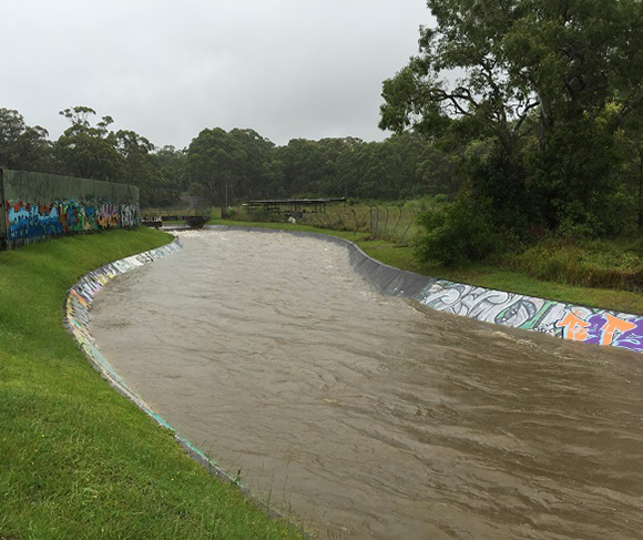 Fast flowing brown stormwater fills a concrete-sided stream with green grass and trees on either side.