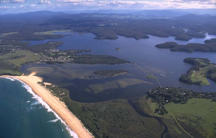 An aerial view of Wallaga Lake, featuring a large body of water with multiple inlets and surrounded by lush greenery. The lake has small islands and peninsulas, with a sandy beach on the left separating it from the ocean. The contrast between the dark blue ocean and the lighter blue-green lake water is striking, with forested areas and open grasslands in the surrounding landscape.