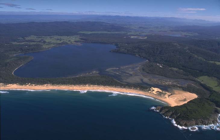 An aerial view of Wallagoot Lake, showcasing the expansive body of water bordered by a dense forest on one side and a curved sandy beach on the other. The lake is calm, while waves from the adjacent ocean crash onto the shore. The scene is set under a clear blue sky.