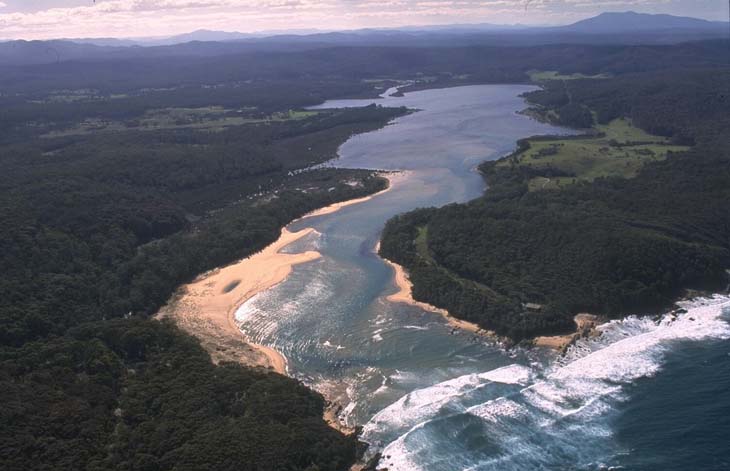 Aerial view of Wapengo Lagoon with a narrow channel connecting it to the ocean. The lagoon is surrounded by dense greenery and a sandy beach. Waves are seen breaking on the shoreline, indicating the meeting point of the lagoon and the sea.