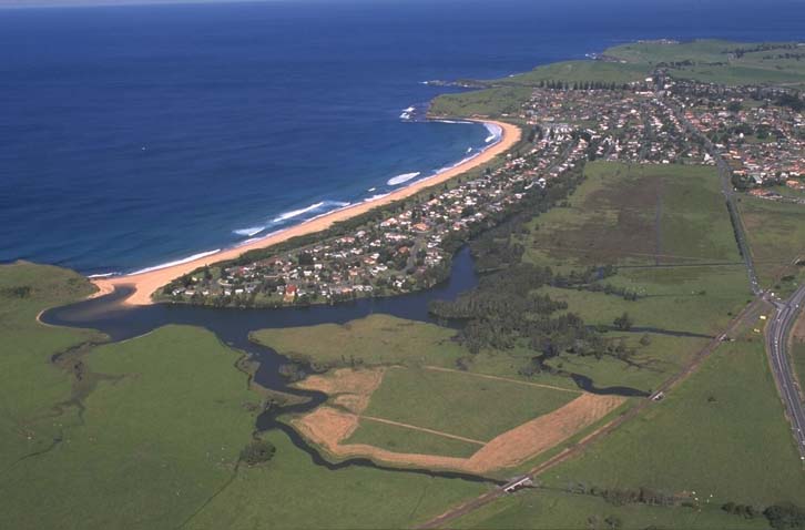 Aerial view showing river running through large green cleared pastures, with a town between the river and the beach. 