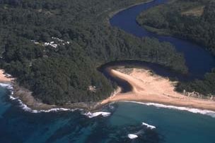 Aerial view of Willinga Lake near the coastline, showing a winding body of water with a narrow point leading into the ocean. Waves crash onto the sandy shore, surrounded by dense greenery and some clearings with buildings amidst the trees, illustrating a blend of natural beauty and human habitation.