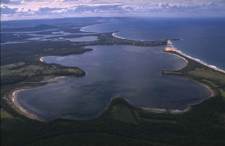 Aerial view of Wollumboola Lake, a coastal lagoon with dark blue waters, surrounded by green vegetation and sandy shores. The lake is near the ocean, separated by a narrow strip of land with visible waves on one side and calm water on the other.