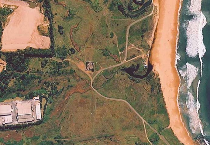 Aerial view of a coastal landscape showing Woodlands Creek flowing into the ocean, with surrounding greenery, trails, and a building complex near the shoreline.