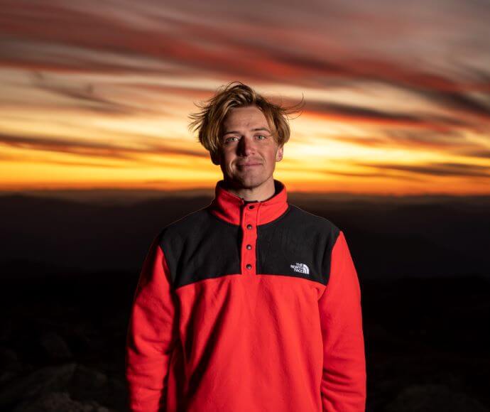 Boen is standing looking at the camera while the sun sets over the mountains in the distance