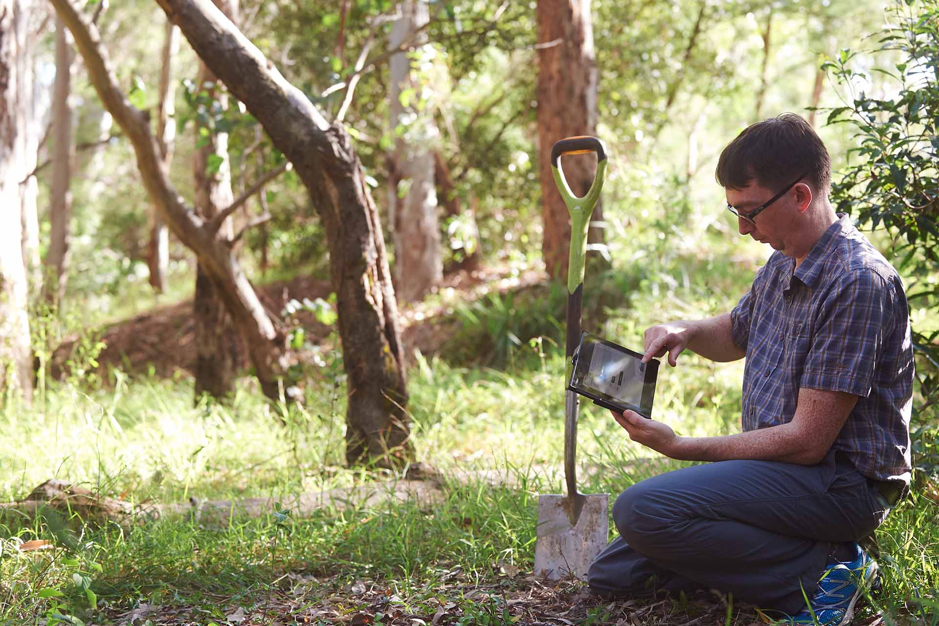 A person is kneeling in a forested area using a tablet next to an upright shovel