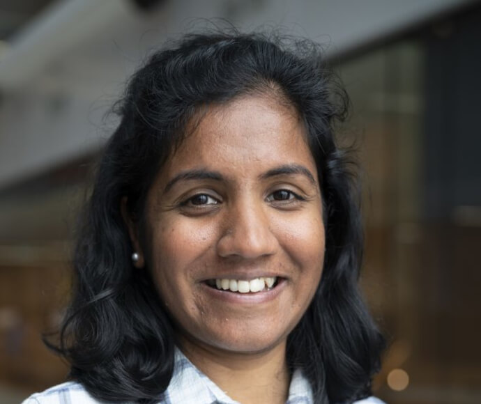 Portrait of a professional woman in a lab coat, smiling confidently. She is standing in a laboratory setting with scientific equipment in the background