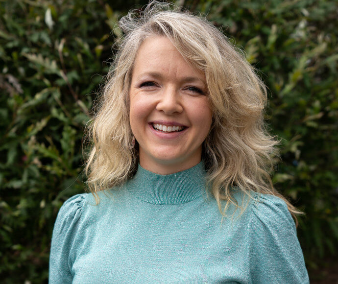 Portrait of a woman, smiling confidently. She is standing in front of a large green bush.. 