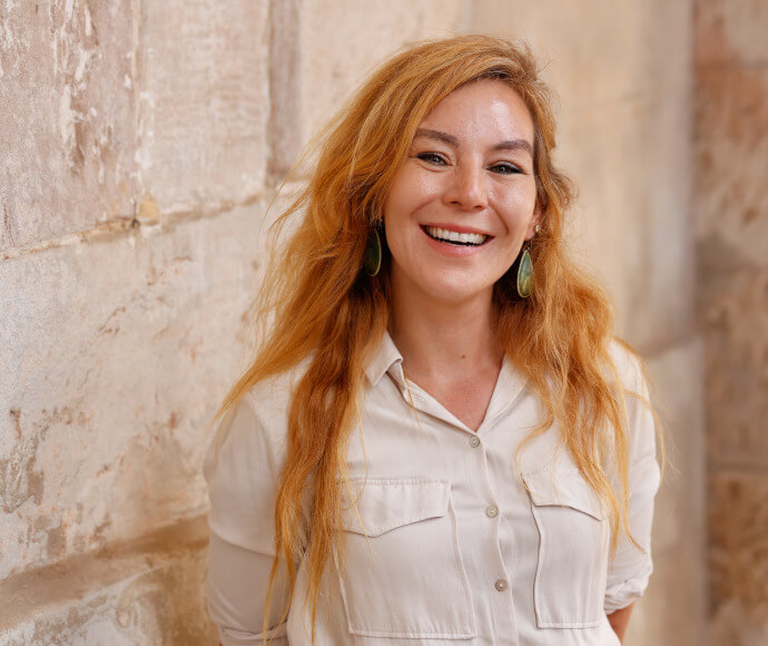 Portrait of a woman with long red-hair, smiling confidently. She is standing front of a stone wall. 