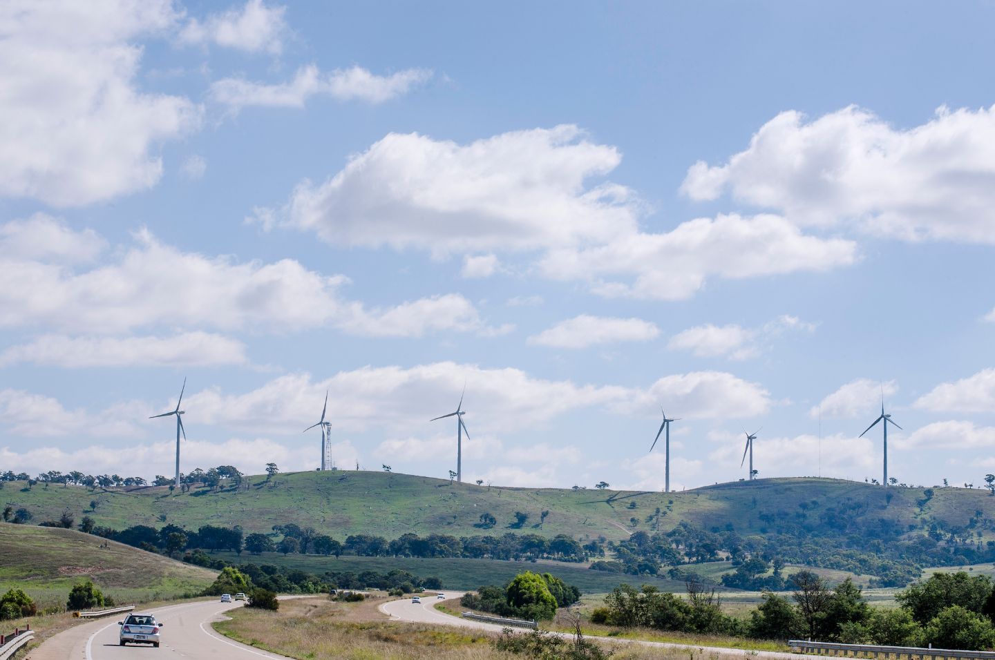 A car moves along a road, framed by the Capital Wind Farm's wind turbines, highlighting the blend of transportation and sustainability.