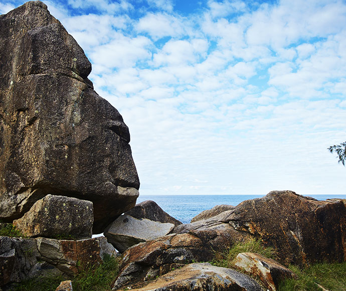 A large rock formation resembling a human face is on the left, surrounded by smaller rocks and grass patches. The ocean is in the background under a partly cloudy sky. This coastal scene likely captures Arakoon National Park, known for beachside camping, rocky foreshores, great fishing, whale watching, and swimming.