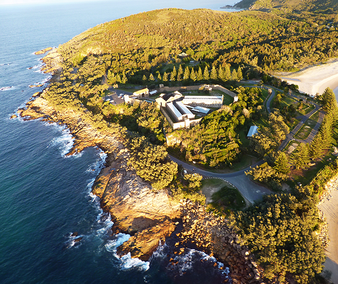 This image shows an aerial view of Trial Bay Gaol campground. The historic gaol building is central, surrounded by greenery and bordered by the ocean and beach. The landscape features winding roads, dense forest, and rocky shorelines. The viewpoint offers a comprehensive look at the natural beauty and historical significance of the coastal location.