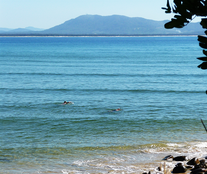 This image shows a serene coastal scene with two people swimming in the ocean. The water is a clear blue-green, with gentle waves visible. In the background, there is a mountain range with a hazy outline, and the sky is clear. The foreground includes a rocky shoreline with some vegetation framing the image on the right side. The image captures a peaceful moment of people enjoying the natural beauty of the ocean and the surrounding landscape.