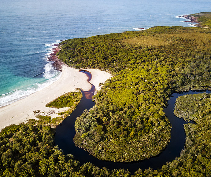 Aerial view of Saltwater campground in Ben Boyd National Park showcasing a coastal landscape with a dense green forest, a winding river meeting the sea, sandy beachfront, and rocky shoreline.