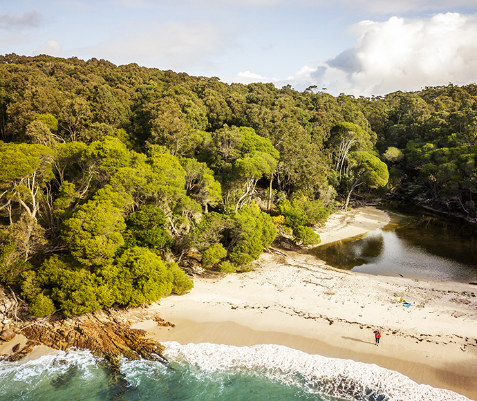 Aerial view of  Bittangabee campground in Ben Boyd National Park showcasing a coastal landscape with a dense green forest, a winding river meeting the sea, sandy beachfront, and rocky shoreline.