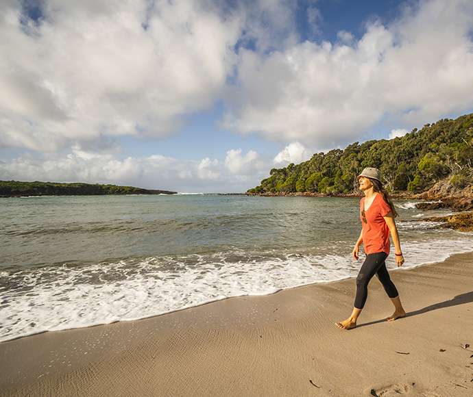 A person walking on a sandy beach beside the ocean, with a backdrop of lush greenery and trees under a partly cloudy sky.