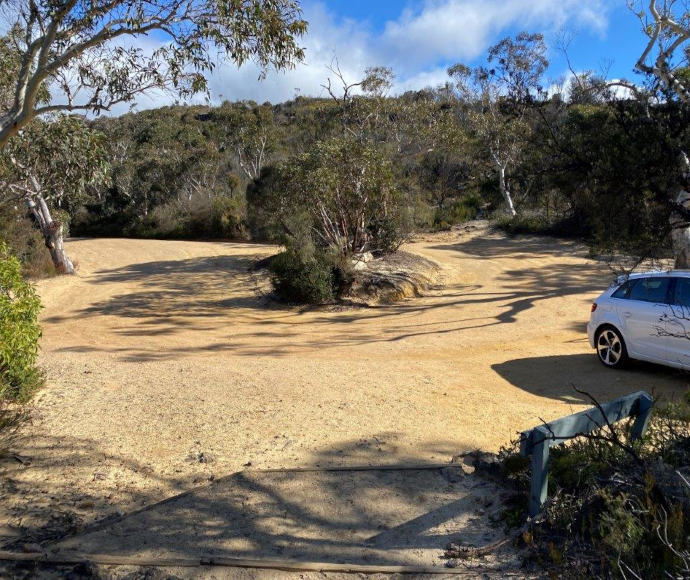 A dirt parking area surrounded by dense greenery under a clear sky. A white car is parked on the left side, and there’s a wooden barrier partially visible in the foreground.