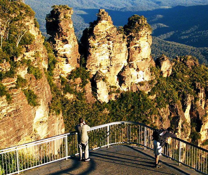 Scenic viewpoint from Echo Point lookout, featuring the Three Sisters rock formation in Blue Mountains National Park. Two individuals stand on a viewing platform, one taking a photo and the other looking out towards the Three Sisters, illuminated by sunlight against a backdrop of dense eucalyptus forests and deep valleys.