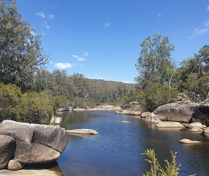 A tranquil river landscape with still water surrounded by large boulders and green trees under a clear blue sky.