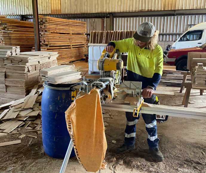 Someone in fluorescent work gear and a cap that says "crusty demons" stands cutting wood; there are large stacks of slats of different shapes behind him in a large corrugated iron shed