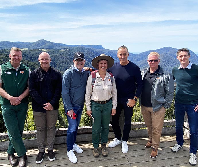 A group photo of stakeholders standing together in front of a lush, green forest backdrop, all smiling and dressed in casual outdoor attire.