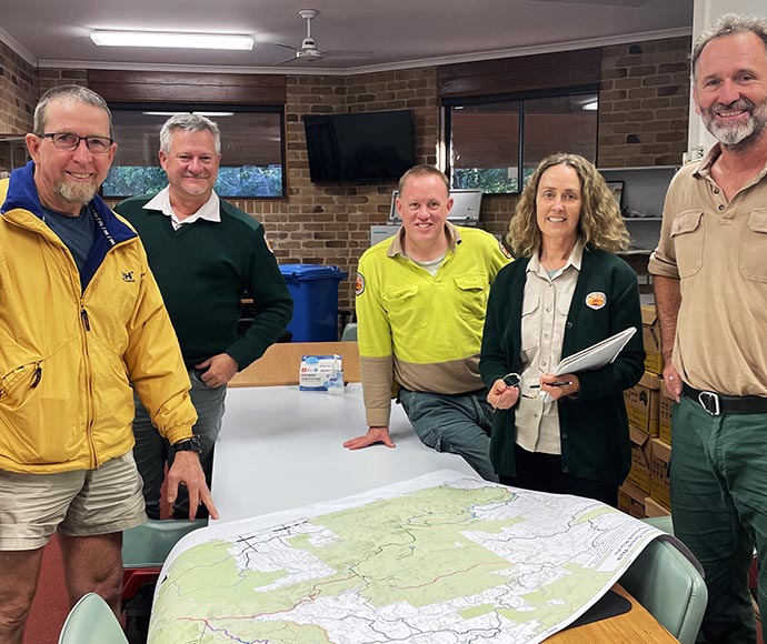 Australian adventurer Peter Treseder AO and four NSW National Parks and Wildlife Service staff members stand indoors with large map on a table, smiling at the camera.