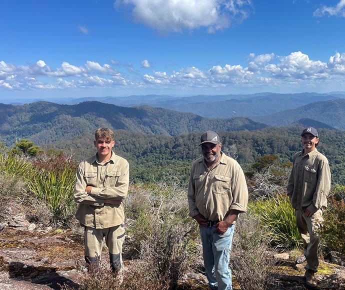 Group photo of Malakai, Philip and Josh from Dorrigo Aboriginal Land Council, dressed in outdoor gear, standing in front of a backdrop of mountains on a bright sunny day with scattered clouds. 