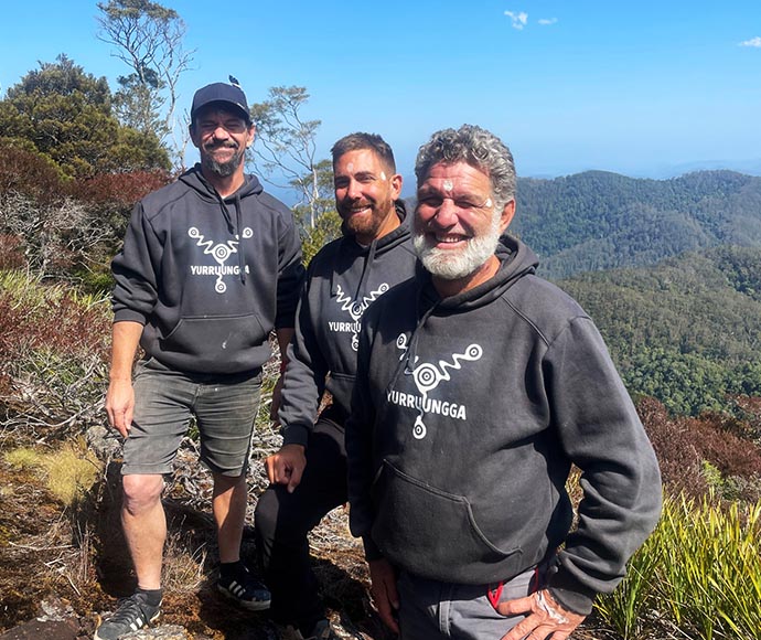 Group photo of Brentyn, Dean and Ciaron from Yurruungga Aboriginal Corporation standing together in front of a backdrop of mountains, all smiling and wearing a black Yurruungga hoodie.