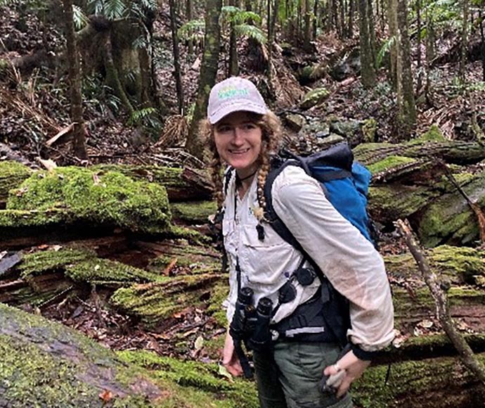 ELA ecologist Phoebe Smith standing in a natural setting within Dorrigo National Park, wearing outdoor field clothing, a hat, binoculars around her neck, and a backpack, surrounded by lush vegetation as she conducts fieldwork.