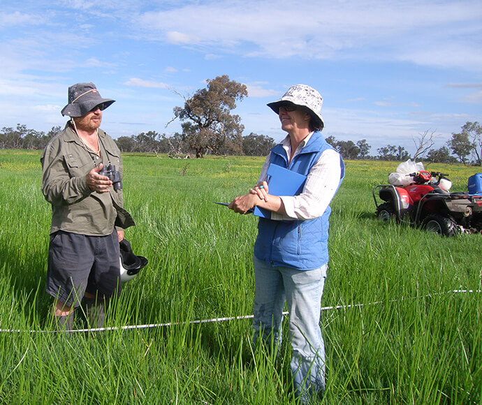 Two individuals from Gwydir EWAG conducting fieldwork on Old Dromana. They are standing in a lush green field with measuring equipment. One person is holding a clipboard and pen, while the other is gesturing towards the ground. An all-terrain vehicle is parked in the background.