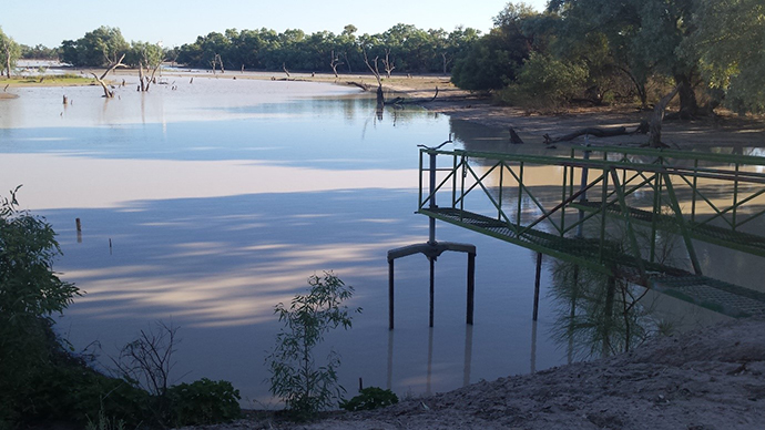 Boera Dam at Toorale, featuring calm water with reflections, surrounded by sparse vegetation and trees. A metal walkway extends over the water on the right side.