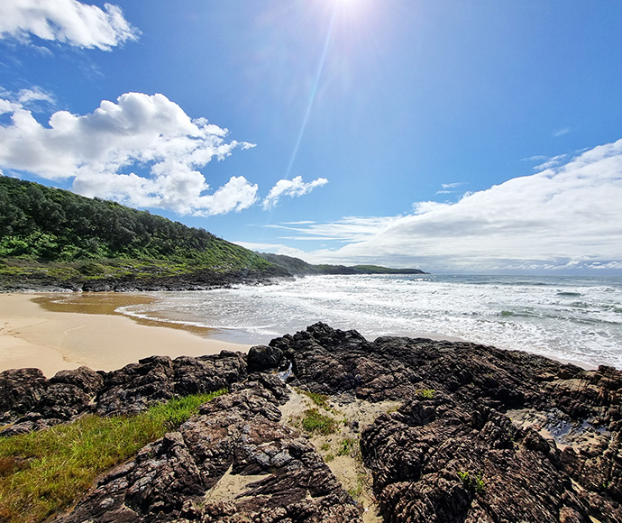 Sunny beach with rocks, waves, and blue sky with clouds.