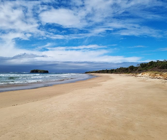 Sandy beach with waves, an island offshore, blue sky with clouds, and a forested coastline.