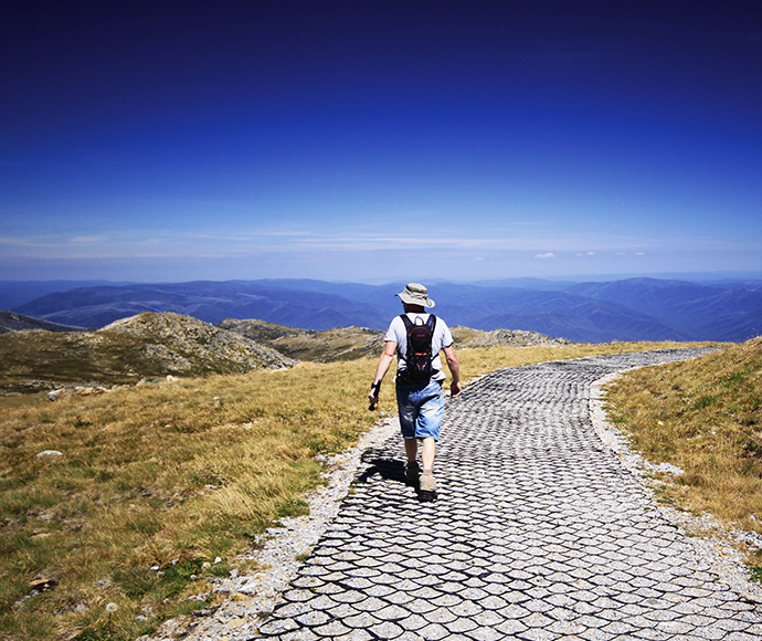 View of the walking track at Kosciuszko National Park, with Mount Kosciuszko in the background. The trail is surrounded by alpine vegetation and offers stunning panoramic views of the rugged landscape.