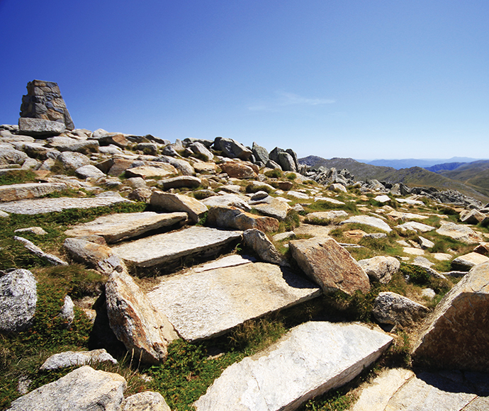 View of the walking track at Kosciuszko National Park, with Mount Kosciuszko in the background. The trail is surrounded by alpine vegetation and offers stunning panoramic views of the rugged landscape.