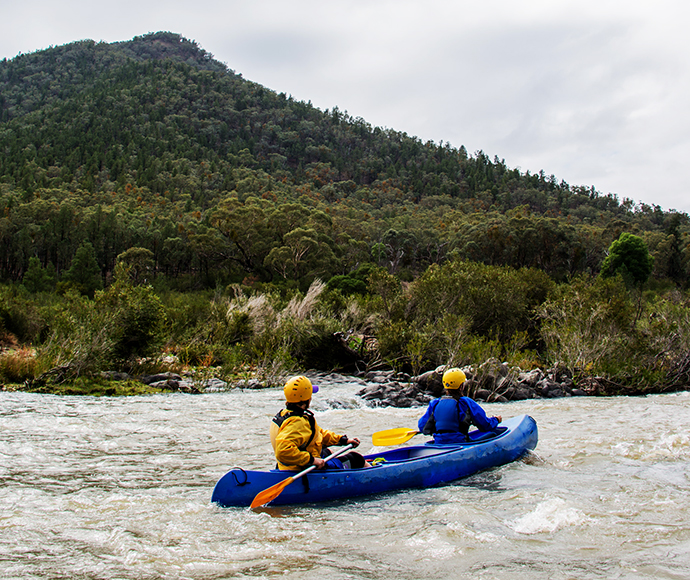 Volunteers paddling a canoe on a serene lake in Kosciuszko National Park. The scene features calm waters surrounded by lush greenery and distant mountains, highlighting the park’s natural beauty and opportunities for outdoor activities.