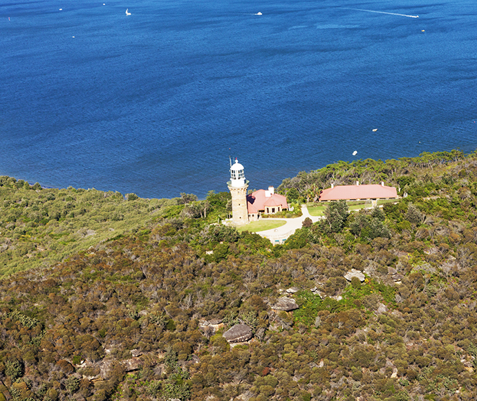 An aerial view of Ku-ring-gai Chase National Park featuring the Barrenjoey Lighthouse at Barrenjoey Head, located at Palm Beach on Sydney’s northern beaches. The top half of the image showcases the clear blue waters of Palm Beach with several boats, while the middle section highlights the lighthouse and surrounding buildings, all enveloped by lush greenery.