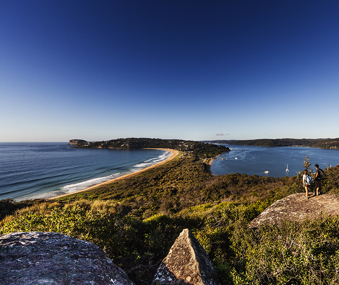 Couple of walkers standing on a rocky outcrop along the Barrenjoey Lighthouse Track, overlooking Palm Beach. The scene captures the stunning coastal views, with the ocean stretching out below and the lush landscape surrounding the trail.