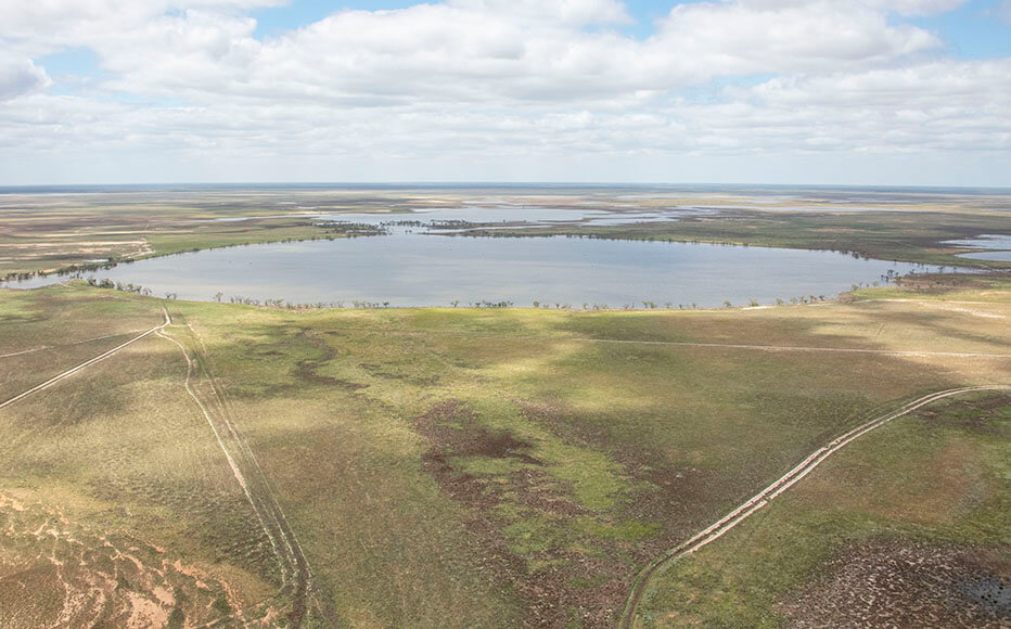 An aerial view of a large body of water surrounded by vast open land. The landscape features a mix of green and brown hues, indicating vegetation and possibly agricultural fields. There are no visible structures or people, emphasizing the natural and rural character of the area. The water reflects the partly cloudy sky above, with sunlight breaking through and casting soft shadows on the ground.