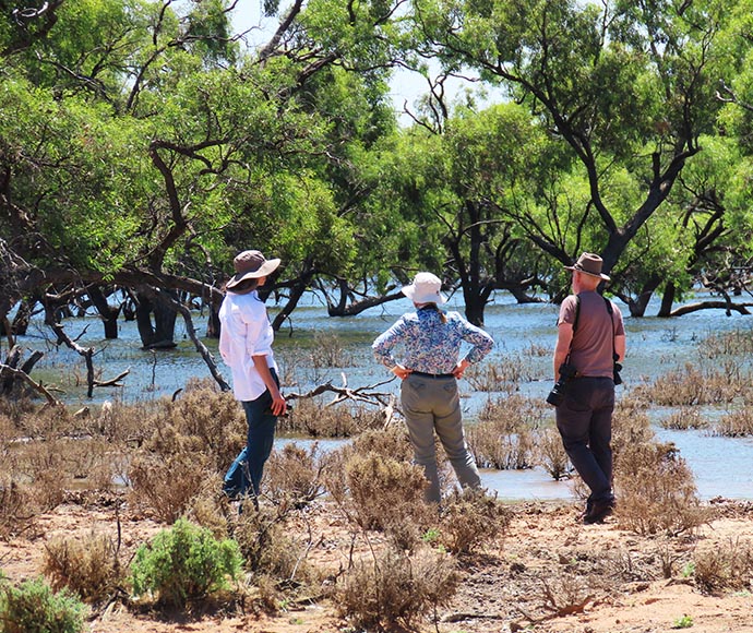 Three individuals stand in a natural setting with water and trees, observing their surroundings. The scene highlights the beauty of nature and the importance of environmental exploration.