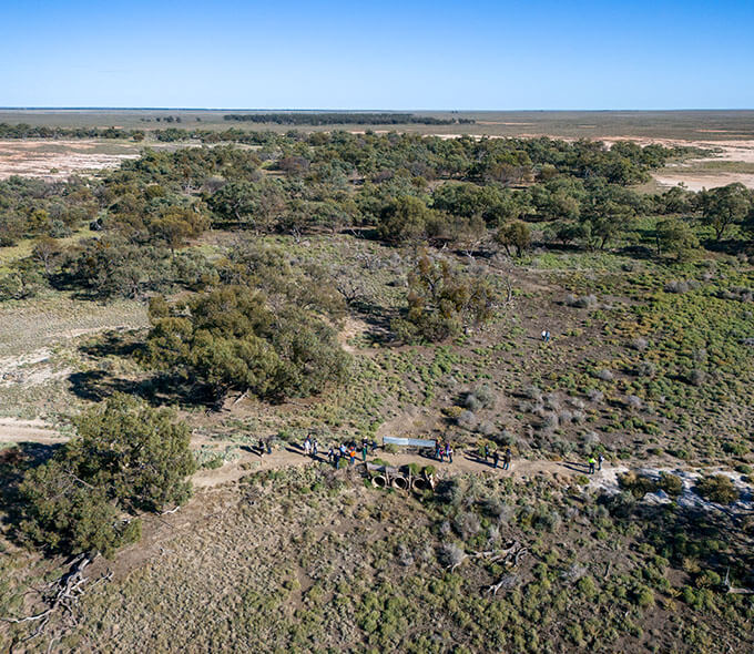 Aerial view of a stark contrast between lush greenery and barren land with a group of people and a vehicle on a path through the vegetated area.