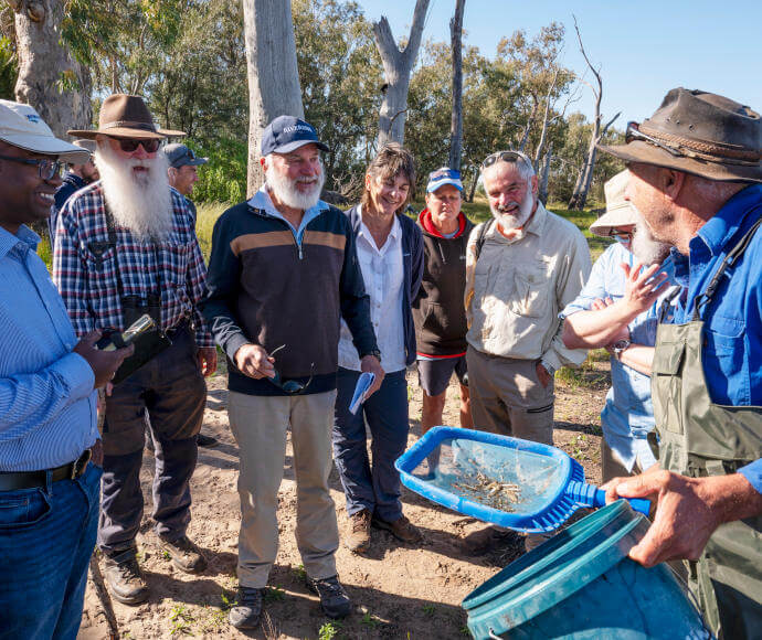 Group of individuals outdoors engaged in an environmental or agricultural activity, with one person holding a blue tray containing soil or organic material, set in a grassy area with trees.