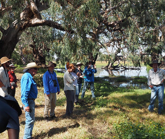 A group of people standing on a dirt road in a rural setting, with fields on either side under a clear blue sky. The group appears to be having an informal meeting or gathering, with some individuals facing each other. Parked vehicles are visible, indicating they traveled to this location.