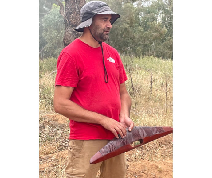 The Bangerang nation's Roland Atkinson talks about the construction and use of tools at the Murray and Lower Darling EWAG meeting. Roland was wearing a red t-shirt and hold a finely carved, wooden Aboriginal artefact with finely engraved patternings.