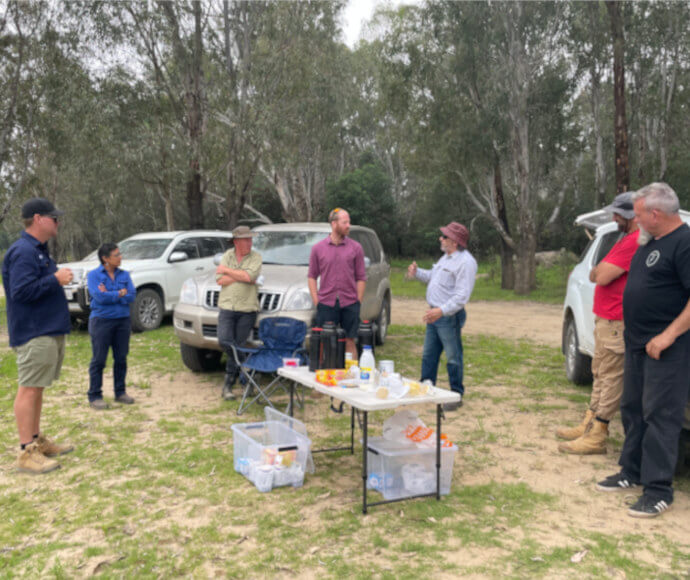 Murray and Lower Darling EWAG members gather around a table with tea things set up outdoors at Buchanan’s Beach, with silvery gumtrees behind them.