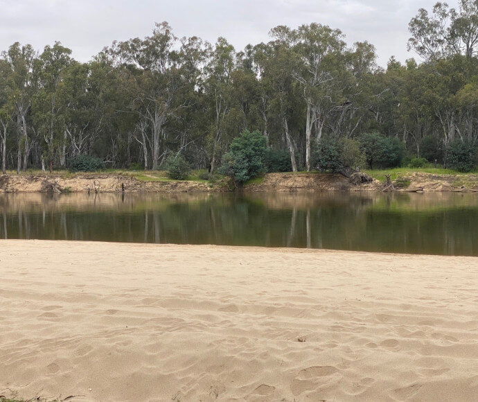 A sandy river shore of Buchanans Beach with still water reflecting trees and grass on the opposite bank.