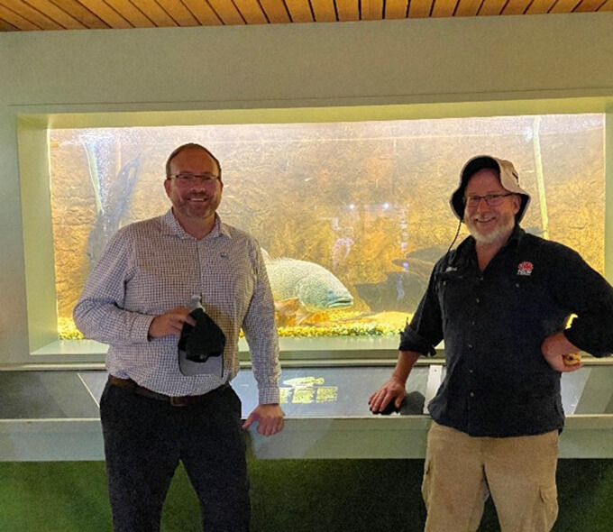 Two individuals standing in front of a large aquarium tank with various fish and aquatic plants visible inside.