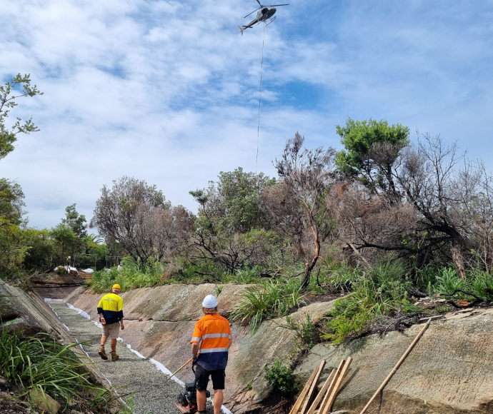 2 workmen in fluoro shirts stand on a narrow path of gravel laid into the floor of an old military ditch, with small windblown trees in the background