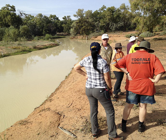 Members of the Toorale Joint Management Committee discussing a project outdoors near a water body with vegetation in the background. Individuals are seen standing on the bank, observing and pointing towards the water, indicating an active engagement with the environmental setting.