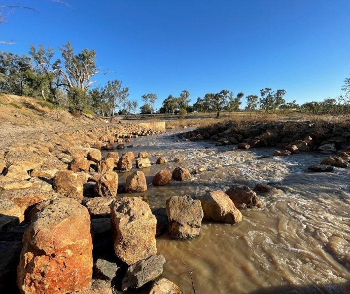 Close-up image of brown river water passing through rock fish traps, with the bush and clear blue sky in the background